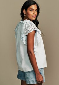 Top in cotone azzurro chiaro con rouches in pizzo ricamato sulle spalle, abbinato a una vestibilità comoda e un tessuto testurizzato, indossato con pantaloni shorts in denim.