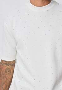 T-shirt blanc à manches courtes en tissu doux, orné de strass en argent dans un motif éparpillé. Présente un col rond.