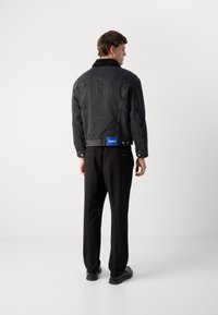 Chaqueta de mezclilla oscura con un cuello de borreguito, con una etiqueta con la marca en azul en la parte trasera, combinada con pantalones negros y zapatos negros.