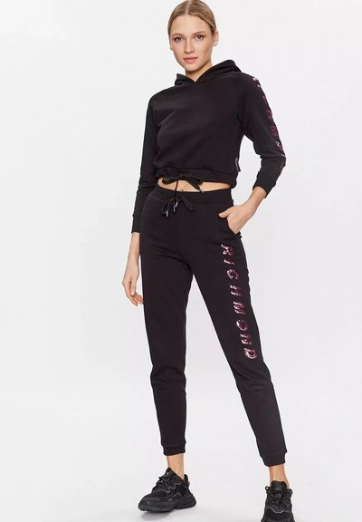 Donna che indossa un cropped hoodie nero e joggers con testo "RICHMOND" in paillettes rosa, in piedi con una mano in tasca e sneakers nere.