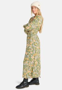 Vestido maxi floral en verde con flores amarillas y moradas; mangas largas, diseño con volantes, combinado con botas de combate negras y un gorro de punto crema.