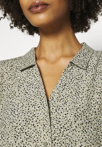 Blusa con cuello en un tejido verde claro con un estampado disperso en blanco y negro, que presenta un cierre de botones y una textura suave.