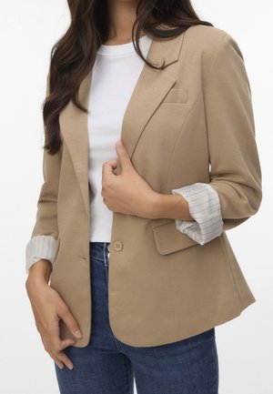 Femme portant un blazer beige avec des poignets rayés retroussés sur une chemise blanche et un jean bleu, tenant le revers du blazer d'une main.