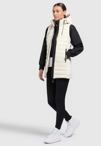 Gepolsterte cremefarbene Weste mit hohem Kragen und Kapuze, kombiniert mit einem schwarzen Langarmoberteil und schwarzen Leggings, abgerundet mit weißen High-Top-Sneakern.