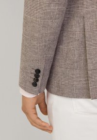 Manche de blazer à motif check marron avec quatre boutons noirs. Poignet de chemise rayée rose clair et blanche visible en dessous. Pantalon blanc en arrière-plan.