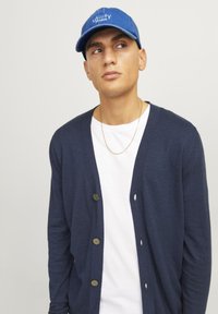 Mørkeblå cardigan med fem brune knapper, b�året over en hvid t-shirt. Personen har en blå kasket på med teksten "utility studios".