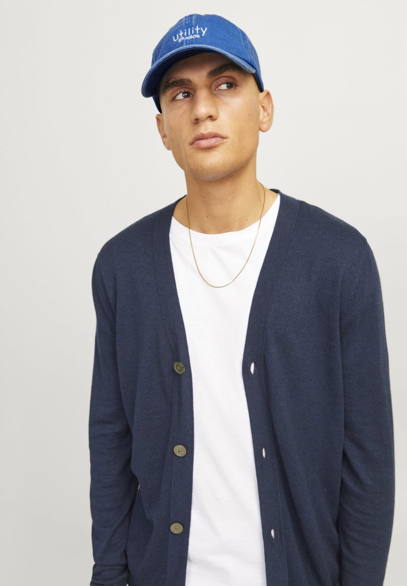 Navyblauer Cardigan mit fünf braunen Knöpfen, getragen über einem weißen T-Shirt. Die Person trägt eine blaue Mütze mit dem Text "utility studios".
