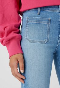 Úzké modré džíny s vybledlou texturou, vysokým pasem a jedinou zadní kapsou. Kombinovány s výraznou růžovou mikinou s žebrovanými rukávy.