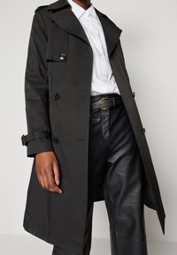 Mörkgrön trenchcoat med dubbelknäppt design, prydd med svarta knappar, stora slag och manschettremmar, bärs över svarta skinnbyxor.