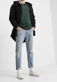 Abrigo largo negro sobre un sudadera verde, combinado con jeans claros desgastados y zapatillas blancas. Calcetines lisos visibles por encima de las zapatillas.