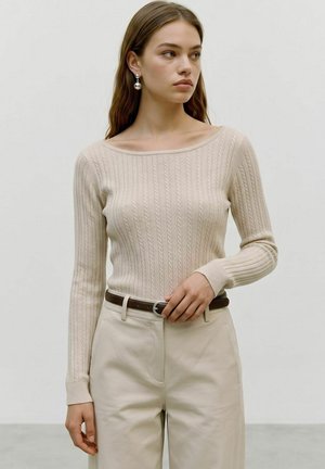Jeune femme aux longs cheveux bruns portant un pull beige en maille torsadée, un pantalon crème, une ceinture marron et des boucles d'oreilles boules argentées regardant vers la droite.
