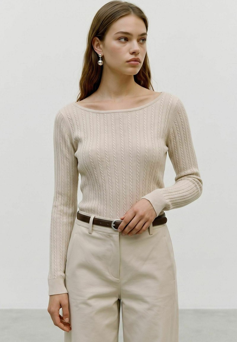 Jeune femme aux longs cheveux bruns portant un pull beige en maille torsadée, un pantalon crème, une ceinture marron et des boucles d'oreilles boules argentées regardant vers la droite.