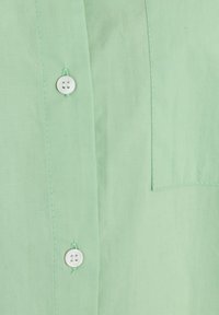 Chemise boutonnée vert clair en tissu lisse, avec une petite poche poitrine et des boutons blancs à deux trous chacun.