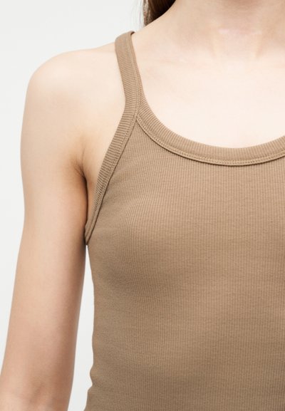 Gros plan sur une personne portant un débardeur beige côtelé, sans manches, avec des bretelles étroites, sur fond blanc.