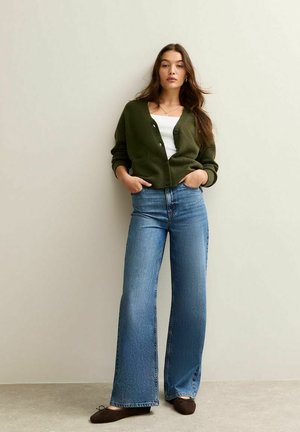 Mörkgrön virkad cardigan med knappar, vit tanktopp, ljusblå vida jeans och bruna platta skor mot en neutral bakgrund.