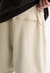 Hand pulling black shirt up to reveal beige textured pants with zippered pocket on the side.