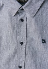 Camicia a maniche corte con bottoni di colore azzurro chiaro, caratterizzata da un motivo testurizzato, bottoni neri e una piccola etichetta con logo sul lato sinistro del petto.