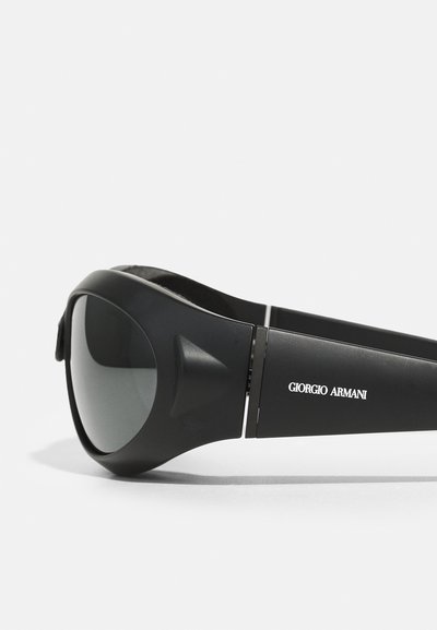Gafas de sol negras con un marco mate, lentes grises y un diseño elegante. Presentan un acento de logo plateado en el lado y una patilla texturizada.