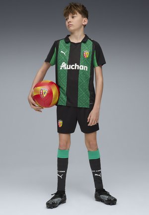 Černý a zelený fotbalový dres s vzorovaným vrškem, šortkami, zelenými akcenty a černými kopačkami. Drží červený a žlutý fotbal.