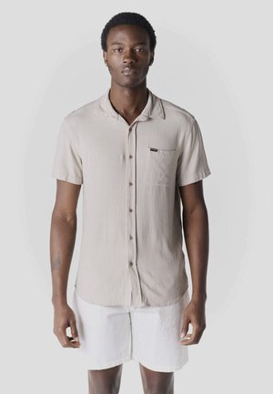 Hombre de pie vistiendo una camisa beige de manga corta con botones y bolsillo, y pantalones cortos blancos, contra un fondo liso y claro.