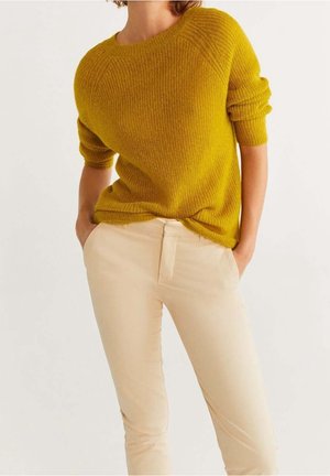 Femme portant un pull côtelé jaune moutarde avec les manches retroussées et un pantalon beige, les mains dans les poches, sur un fond blanc.