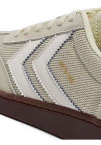 Bež superga s teksturiranim zgornjim delom iz tkanine, belimi usnjenimi poudarki, rjavim gumijastim podplatom in kontrastnimi detajli šivov.