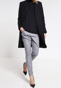 Černý dlouhý kabát, černý rolák, šedé tailored kalhoty a černé lodičky s špičatou špičkou. Hladké textury, přiléhavý design a neutrální barevná paleta.