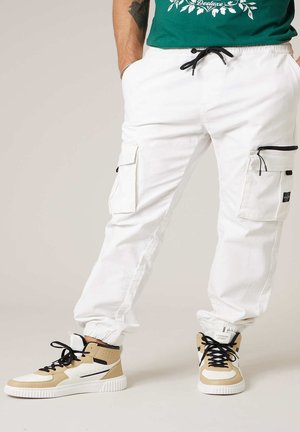 Personne portant un pantalon cargo blanc avec des poches à glissière et à rabat, des baskets montantes beige et blanches avec des lacets noirs, debout sur un fond clair.