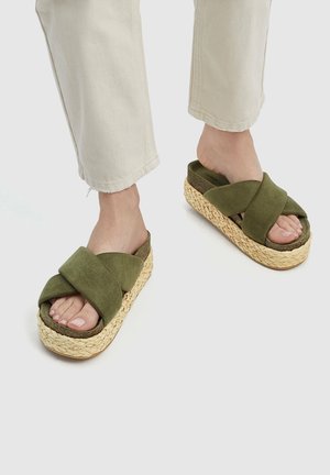 Nohy v béžových kalhotách a olivově zelených platformových sandálech s tkanými slámovými podrážkami na světlém pozadí.