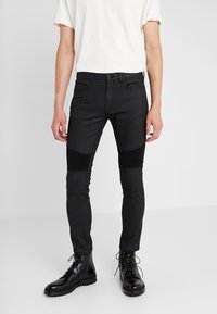 Jeans negros de corte slim con paneles texturizados en las rodillas, bolsillo con cremallera y detalles de costura sutiles. Complementados con botas negras con cordones.