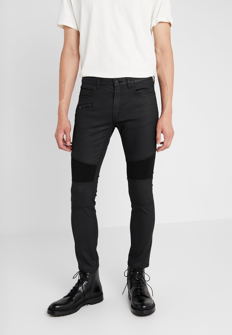 Jeans negros de corte slim con paneles texturizados en las rodillas, bolsillo con cremallera y detalles de costura sutiles. Complementados con botas negras con cordones.