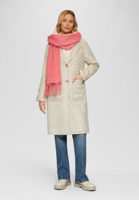 Beige geruite jas met grote zakken en twee knopen, gecombineerd met een roze gebreide sjaal. gedragen over een crème top en blauwe jeans.