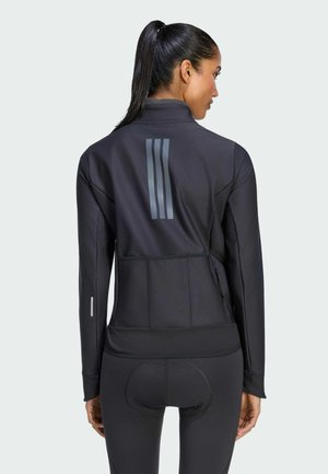 Mujer con coleta oscura que lleva una chaqueta deportiva negra de manga larga y mallas, vista desde atrás sobre un fondo claro.