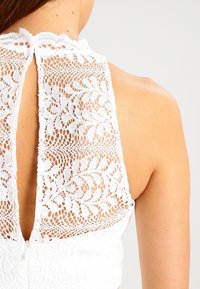 Vit spetsblus med hög halsringning, intrikata blommönster och en nyckelhålsdesign bak. Texturen är delikat och luftig.