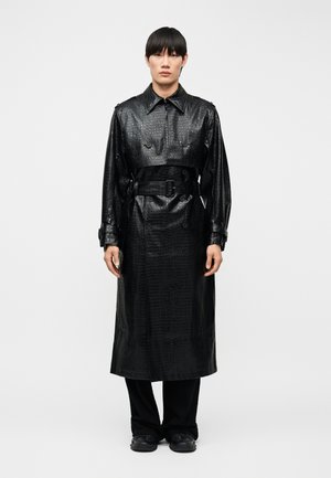 Junge Person trägt einen langen schwarzen Trenchcoat aus künstlichem Krokodilleder mit Gürtel und steht aufrecht vor einem schlichten weißen Hintergrund.