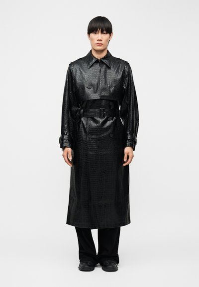 Jeune personne portant un trench-coat long en similicuir noir effet crocodile avec ceinture, debout droit devant un fond blanc uni.