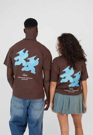 Dwie osoby stoją tyłem, ubrane w brązowe koszulki z dużymi niebieskimi postaciami z kreskówek oraz białym napisem "Legacy" na dolnej części pleców.