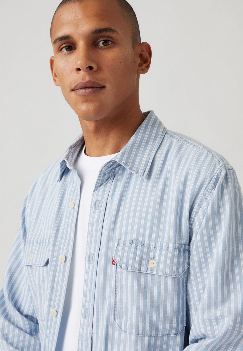 Hellblau gestreiftes Button-up-Hemd aus Baumwolle mit zwei vorderen Taschen, einem Kragen und einem kleinen roten Logo-Detail.