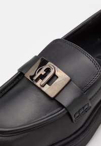 Mocassin en cuir noir avec une boucle métallique argentée présentant un logo "F" stylisé et des coutures détaillées sur la partie supérieure.