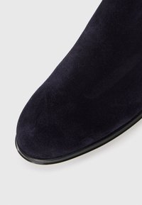 Close-up de um bico de bota de camurça preta com textura suave e uma borda fina de sola de borracha preta contra um fundo branco.