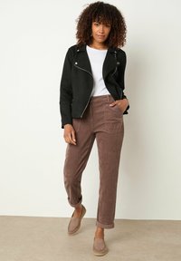 Giacca da moto in suede nera, t-shirt bianca, pantaloni in velluto a coste marroni con tasche laterali e scarpe slip-on chiare. Capelli ricci incorniciano il viso.