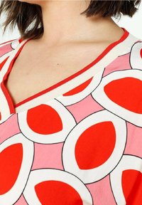 Rød og pink kortærmet top med V-udskæring, prydet med et stort ovalt mønster omrids i sort. Afsluttet med en rød ribbestrikket halsudskæring.