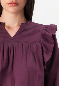 Lähi-kuva naisen kauluksesta, jossa on violetti paita, jossa on röyhelöiset hihat ja koristeellista ompelua, korostaen hienostunutta kaula-aukkoa.