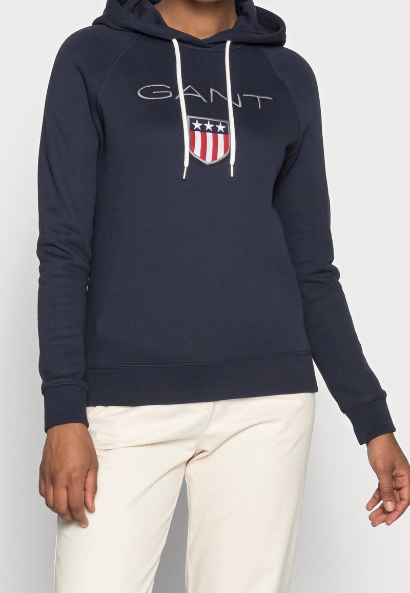 Personne portant un sweat à capuche GANT bleu marine avec des cordons blancs et un logo écusson rouge, blanc et bleu, associé à un pantalon beige clair.