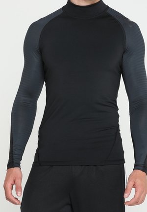 Homme portant un haut de sport noir ajusté à manches longues avec des manches gris foncé et un short de sport noir, debout devant un fond uni.