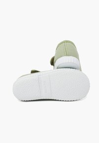 Zapato de lona verde con suela de goma blanca, que presenta una correa acolchada y una suela texturizada. La vista trasera muestra detalles de costura y el logo.