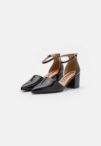 Zapatos de tacón de charol negro con punta afilada, correa al tobillo y tacón cuadrado. El interior presenta una plantilla beige con el logo.