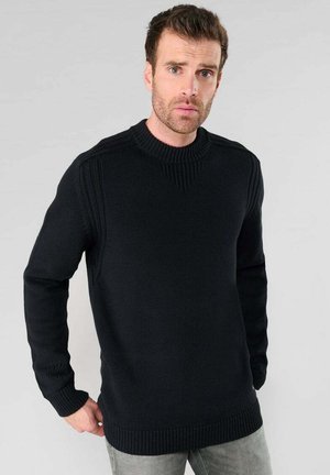 Schwarzer Strickpullover mit langen Ärmeln, geripptem Kragen und strukturiertem Muster; getragen über grauen Jeans, vor einem neutralen Hintergrund gezeigt.