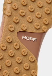 Suela de zapatilla con superficie de caucho texturizado color marrón claro que presenta tachuelas hexagonales y una sección marrón que muestra el nombre de la marca "HOFF" en blanco.