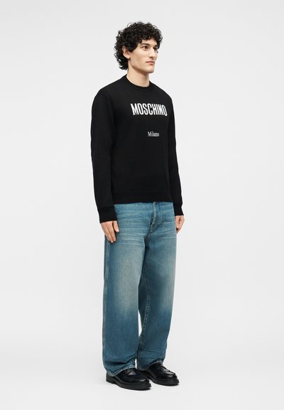Homme debout portant un sweat-shirt noir Moschino, un jean ample bleu clair et des chaussures noires sur un fond blanc uni.